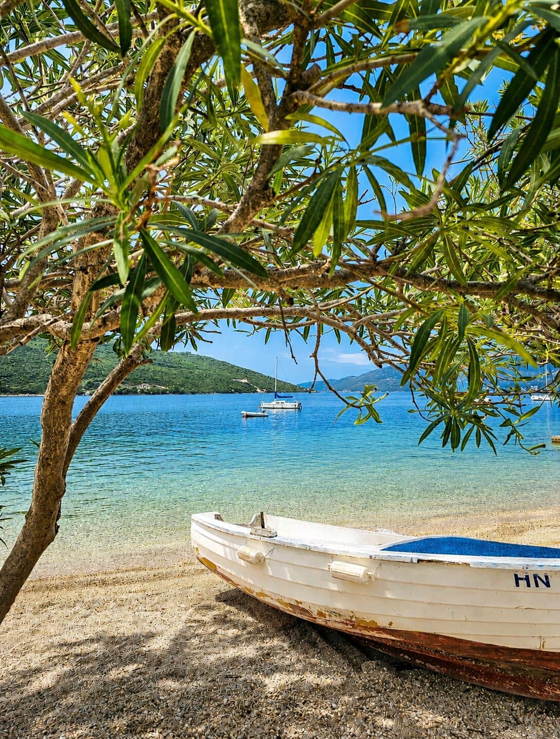 A wooden boat on the pebble shore under the oleander, sailboats in the bay